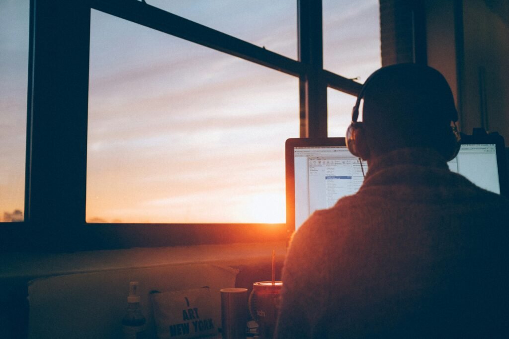 a man sitting at a computer with his back to the camera. behind him is a window with a sun setting